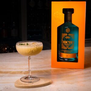 A Bacan Guaro bottle in its sleek orange box next to a crafted cocktail in a coupe glass, garnished with herbs, on a marble bar surface.