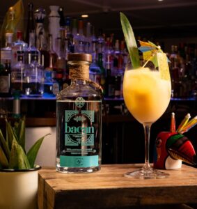 A bottle of Bacan Guaro alongside a tropical cocktail, garnished with pineapple and an umbrella, set on a wooden bar counter with bottles in the background